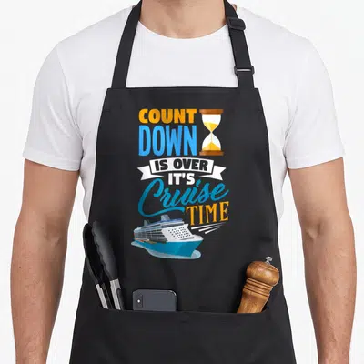 Cruising countdown apron - Stacked orange and blue text with hourglass and blue cruise ship illustration — shown on a black cotton canvas apron against a white background