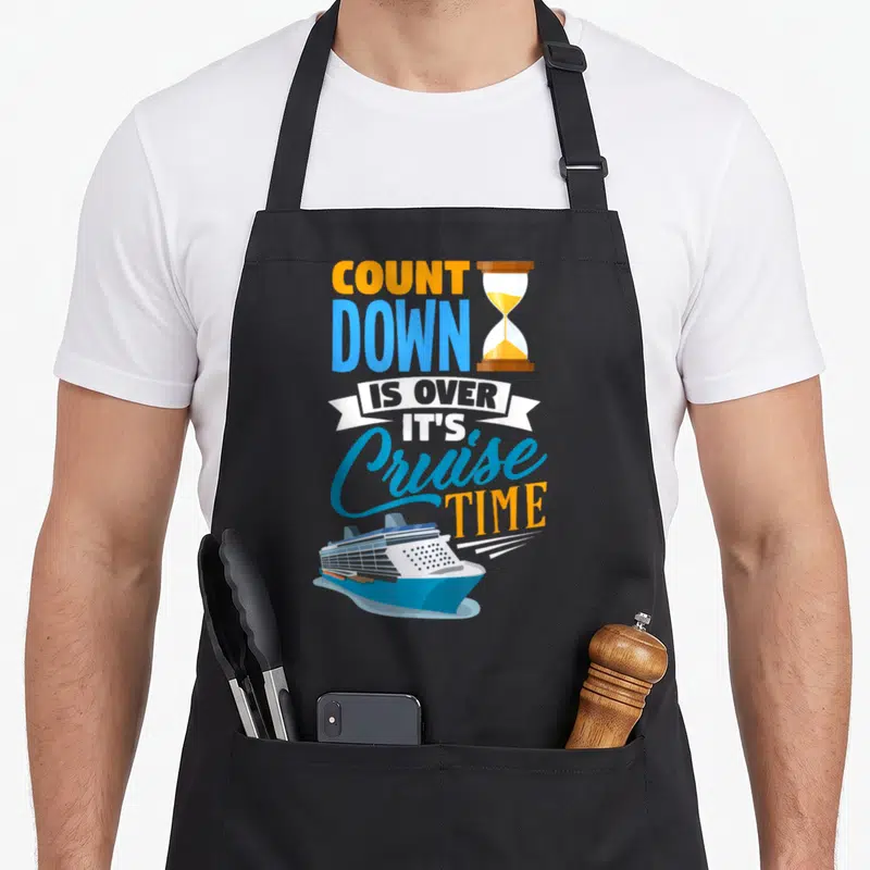 Cruising countdown apron - Stacked orange and blue text with hourglass and blue cruise ship illustration — shown on a black cotton canvas apron against a white background