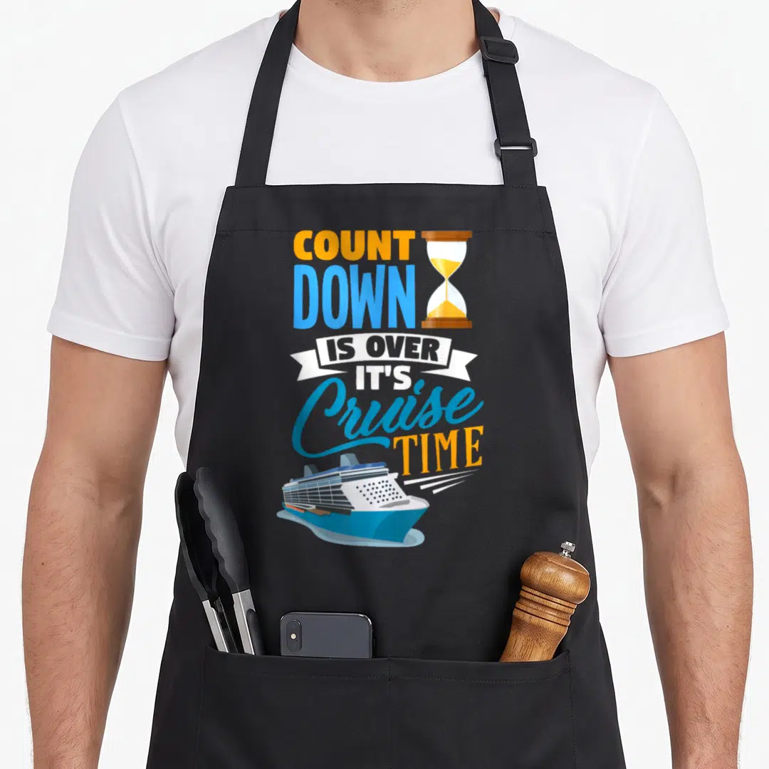 Cruising countdown apron - Stacked orange and blue text with hourglass and blue cruise ship illustration — shown on a black cotton canvas apron against a white background
