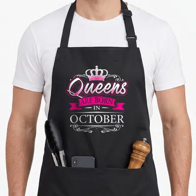 Funny queen october birthday apron - Crown and pink ribbon text reading Queens Are Born In October with white swirls — shown on a black cotton canvas apron against a white background