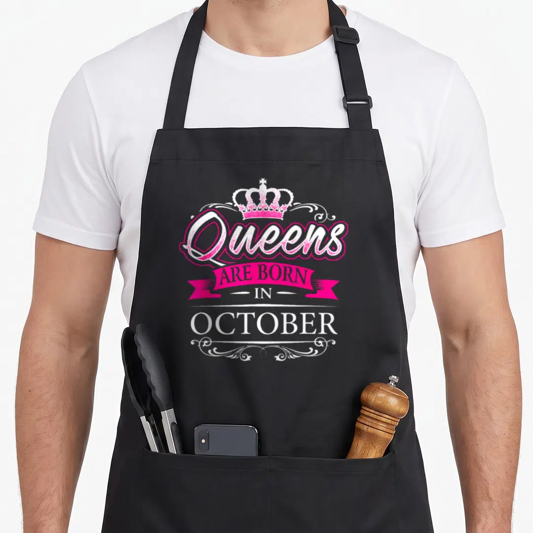 Funny queen october birthday apron - Crown and pink ribbon text reading Queens Are Born In October with white swirls — shown on a black cotton canvas apron against a white background