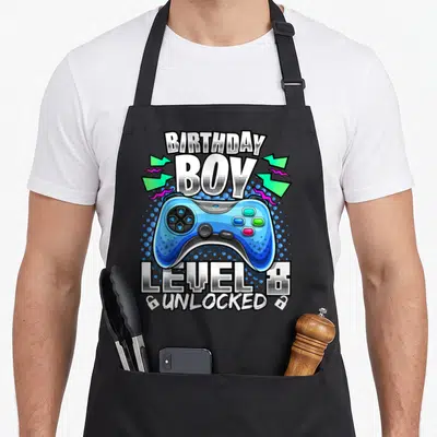 8th video game apron - Blue game controller with BIRTHDAY BOY text and open locks below — shown on a black cotton canvas apron against a white background