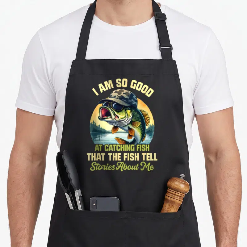 Story fish apron - Cartoon fish wearing hat and sunglasses with arched and stacked text about catching fish — shown on a black cotton canvas apron against a white background
