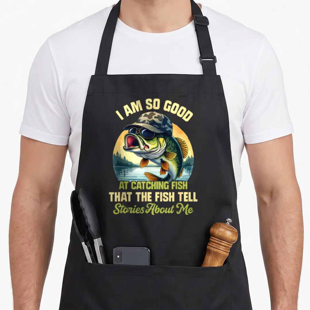 Story fish apron - Cartoon fish wearing hat and sunglasses with arched and stacked text about catching fish — shown on a black cotton canvas apron against a white background