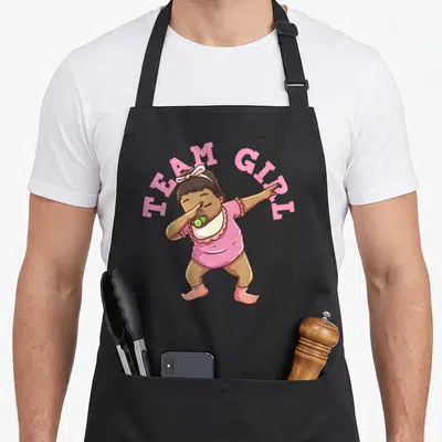 Reveal pregnancy by company apron - Cartoon baby dabbing in pink onesie with arched TEAM GIRL text — shown on a black cotton canvas apron against a white background