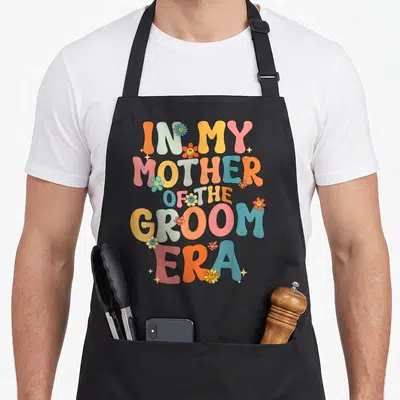 Era mother's apron - Stacked retro text IN MY MOTHER OF THE GROOM ERA with smiley flowers in orange blue pink yellow — shown on a black cotton canvas apron against a white background