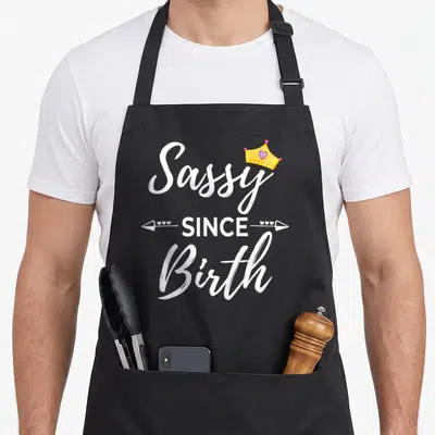 Birth sassy apron - White Sassy SINCE Birth text with yellow crown and arrows — shown on a black cotton canvas apron against a white background