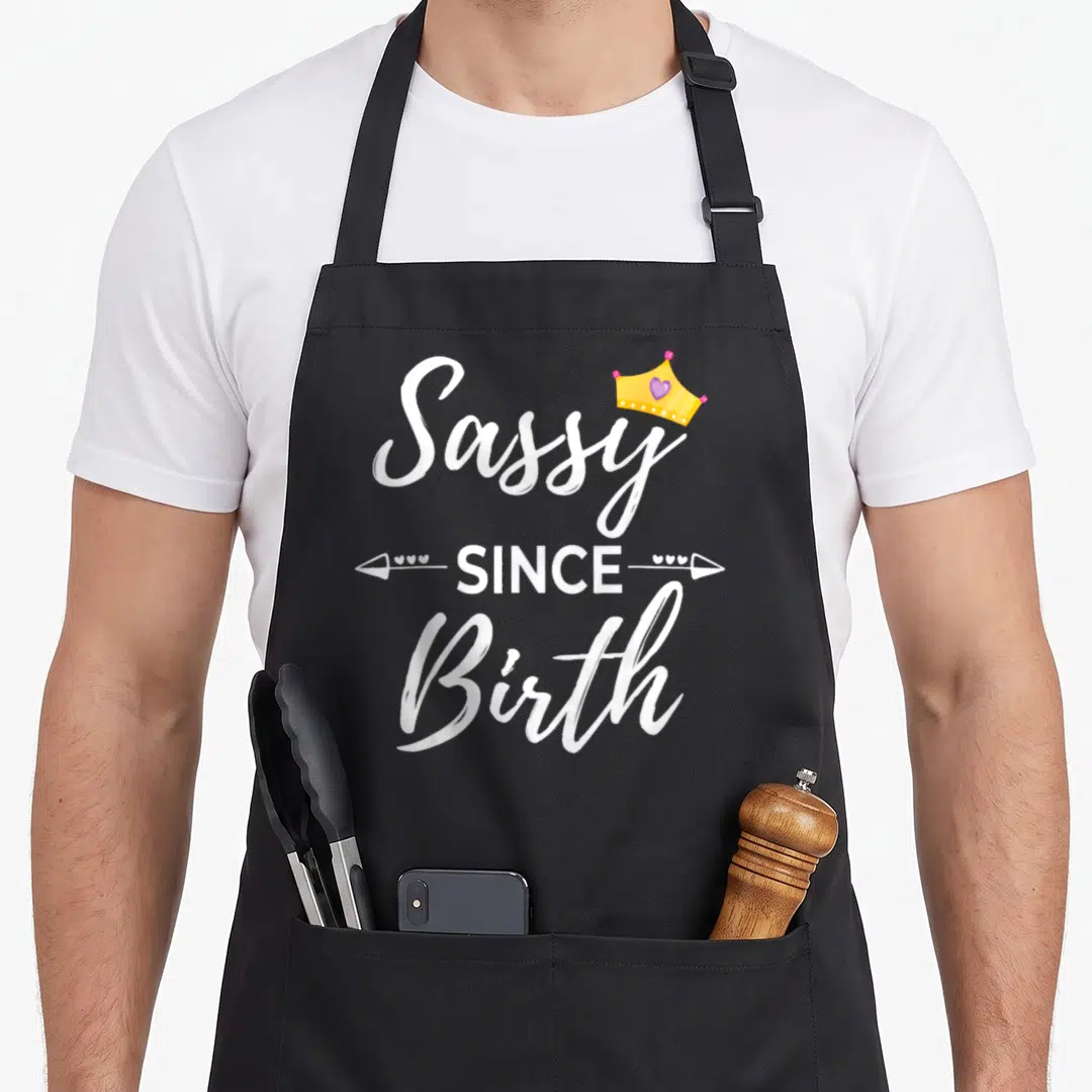 Birth sassy apron - White Sassy SINCE Birth text with yellow crown and arrows — shown on a black cotton canvas apron against a white background