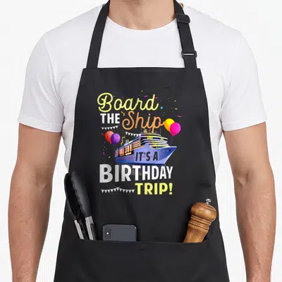 Cruising board birthday apron - Cartoon cruise ship with balloons and text reading Board THE Ship IT'S-A BIRTHDAY TRIP! — shown on a black cotton canvas apron against a white background