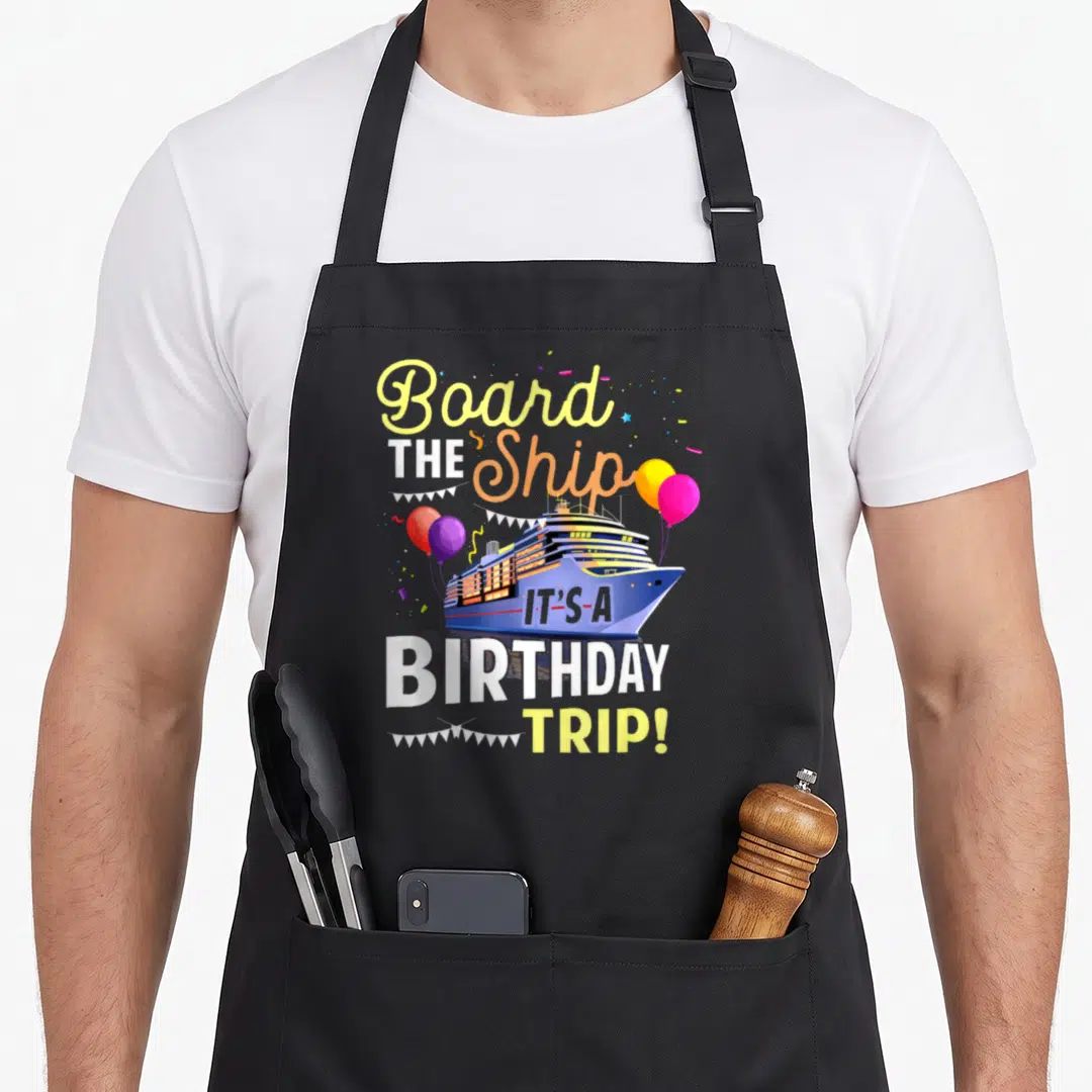 Cruising board birthday apron - Cartoon cruise ship with balloons and text reading Board THE Ship IT'S-A BIRTHDAY TRIP! — shown on a black cotton canvas apron against a white background
