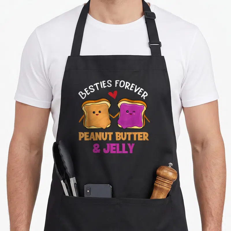 Besty butter apron - Two cartoon toast slices holding hands under Besties Forever text and red heart — shown on a black cotton canvas apron against a white background