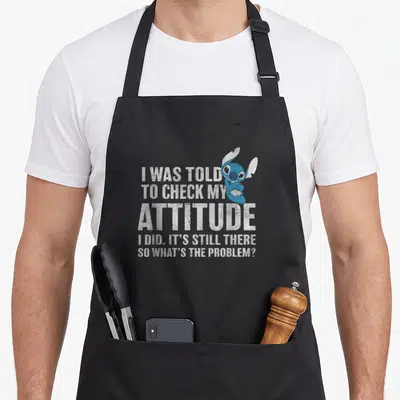 Problem attitude aprons - Stitch character and sarcastic quote about attitude in distressed white and blue text — shown on a black cotton canvas apron against a white background