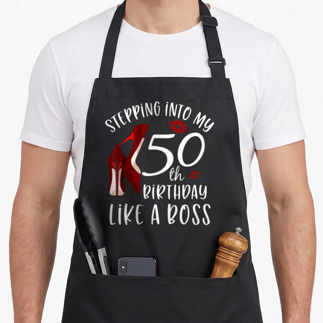 50th bbq boss apron - White STEPPING INTO MY 50th BIRTHDAY LIKE A BOSS text with red glitter high heels and kiss marks — shown on a black cotton canvas apron against a white background