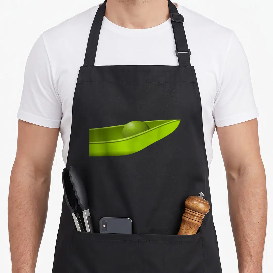 Peas halloween apron - Green cartoon pea sitting inside an open pea pod on black fabric — shown on a black cotton canvas apron against a white background