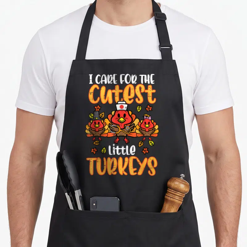 Care thanksgiving apron - Three cartoon turkeys with nurse hat stethoscope and pilgrim hat under text I CARE — shown on a black cotton canvas apron against a white background