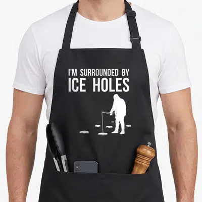 Ice fishing aprons - White text I'M SURROUNDED BY ICE HOLES above fisherman silhouette and oval holes on black — shown on a black cotton canvas apron against a white background