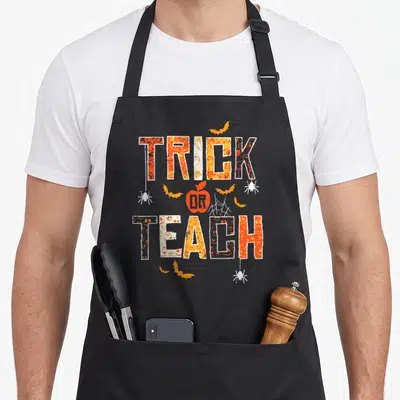 Teach halloween apron - Orange and white Trick Or Teach text with apple and bat graphics — shown on a black cotton canvas apron against a white background