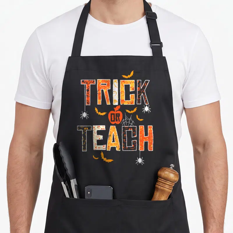 Teach halloween apron - Orange and white Trick Or Teach text with apple and bat graphics — shown on a black cotton canvas apron against a white background