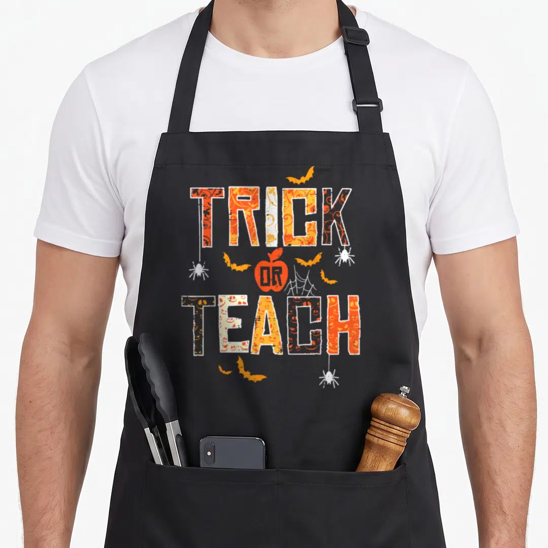 Teach halloween apron - Orange and white Trick Or Teach text with apple and bat graphics — shown on a black cotton canvas apron against a white background