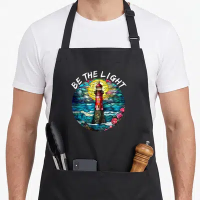 Light stained apron - Stained glass lighthouse with BE THE LIGHT text, blue waves, pink flowers, and yellow sun — shown on a black cotton canvas apron against a white background