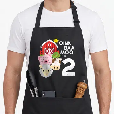 Oink 2nd apron - Red barn, pig, cow, sheep, and OINK BAA MOO I'M 2 text in cartoon style — shown on a black cotton canvas apron against a white background
