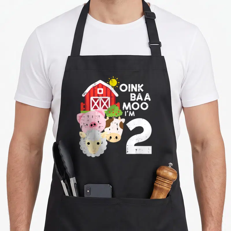 Oink 2nd apron - Red barn, pig, cow, sheep, and OINK BAA MOO I'M 2 text in cartoon style — shown on a black cotton canvas apron against a white background
