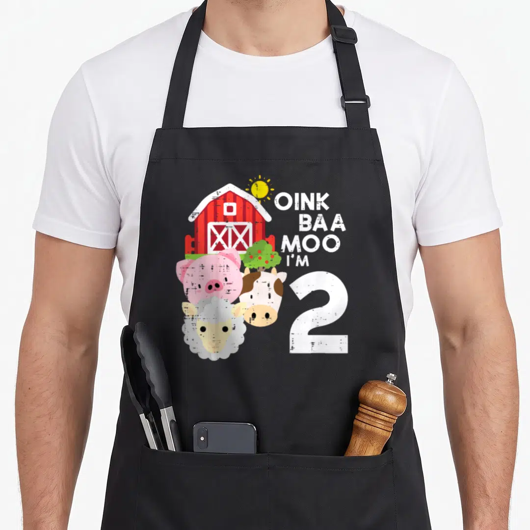 Oink 2nd apron - Red barn, pig, cow, sheep, and OINK BAA MOO I'M 2 text in cartoon style — shown on a black cotton canvas apron against a white background