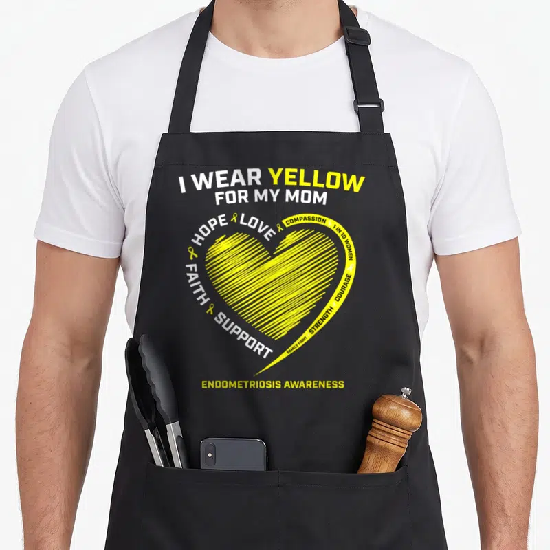 Cute endometriosis awareness apron - I WEAR YELLOW FOR MY MOM text surrounding a yellow striated heart with awareness — shown on a black cotton canvas apron against a white background