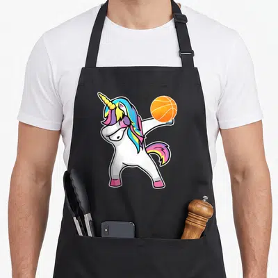 Basketball unicorn apron - Cartoon white unicorn dabbing while holding an orange basketball aloft with rainbow mane — shown on a black cotton canvas apron against a white background