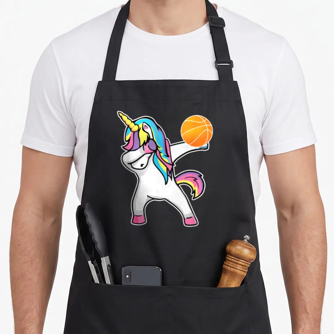Basketball unicorn apron - Cartoon white unicorn dabbing while holding an orange basketball aloft with rainbow mane — shown on a black cotton canvas apron against a white background