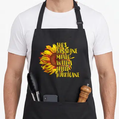 Sunshine mixed hurricane apron - Vertical text SHE'S SUNSHINE MIXED WITH A LITTLE HURRICANE over a yellow orange — shown on a black cotton canvas apron against a white background