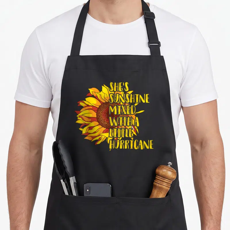 Sunshine mixed hurricane apron - Vertical text SHE'S SUNSHINE MIXED WITH A LITTLE HURRICANE over a yellow orange — shown on a black cotton canvas apron against a white background
