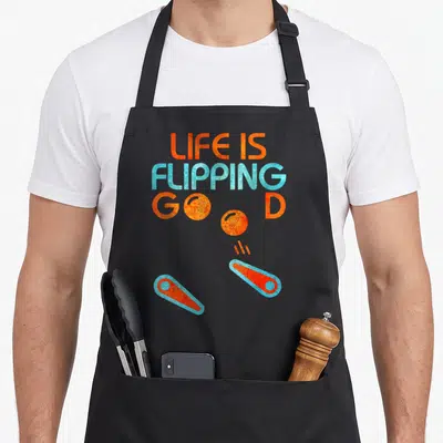 Flipping pinball apron - Orange and blue Life is Flipping Good text with bouncing ball and bumper graphics — shown on a black cotton canvas apron against a white background