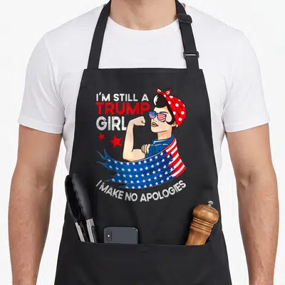 Apology headband apron - Cartoon woman flexing arm with I'M STILL A TRUMP GIRL I MAKE NO APOLOGIES text and three red — shown on a black cotton canvas apron against a white background