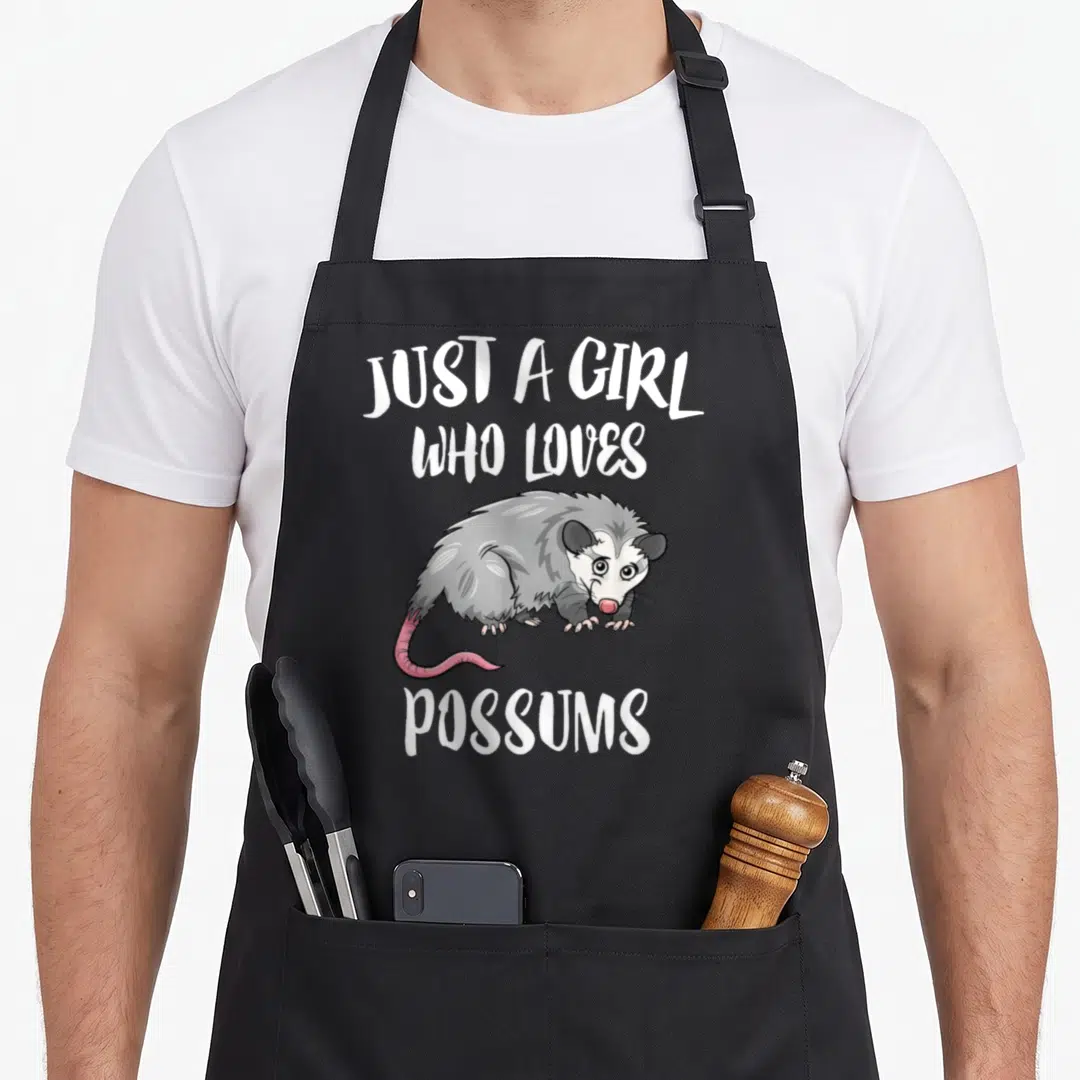 Possum animal apron - Cartoon grey opossum with pink tail and white text Just A Girl Who Loves Possums — shown on a black cotton canvas apron against a white background