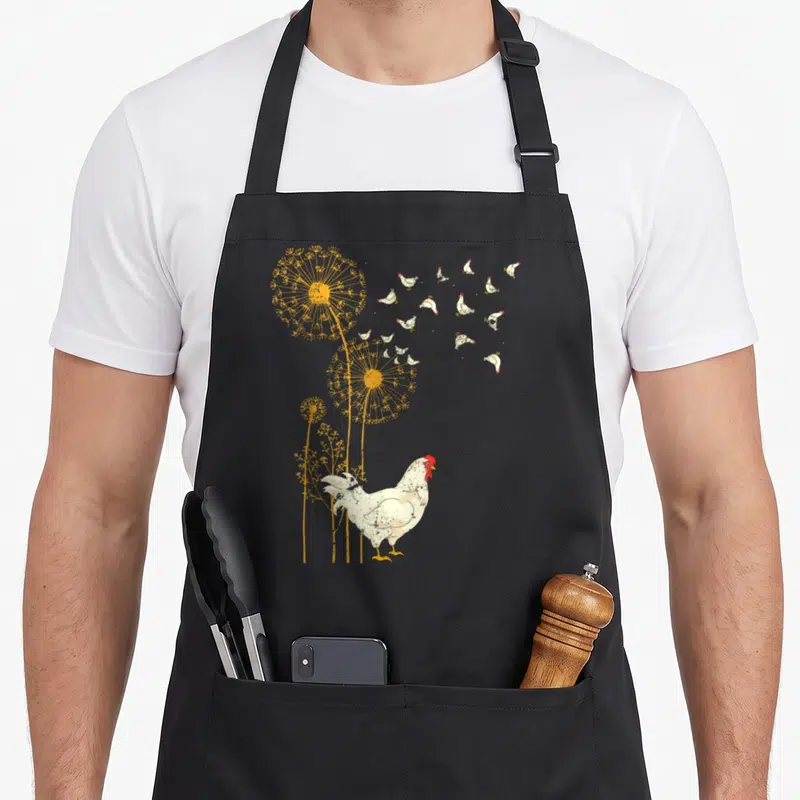 Farmer birds apron - White chicken standing below yellow dandelions with small floating chickens drifting right — shown on a black cotton canvas apron against a white background