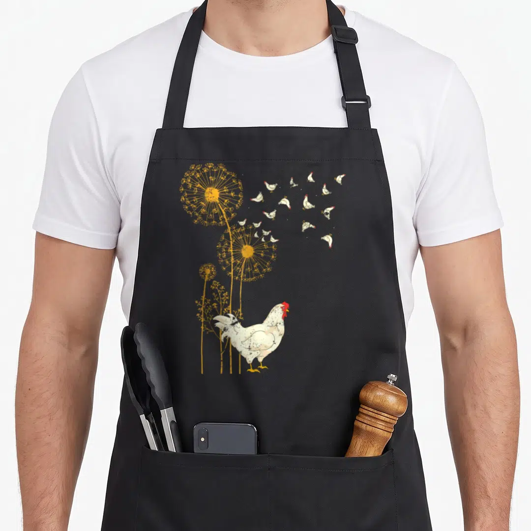 Farmer birds apron - White chicken standing below yellow dandelions with small floating chickens drifting right — shown on a black cotton canvas apron against a white background