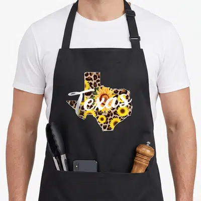 Texas leopard apron - Texas state map with brown leopard print, yellow sunflowers, and white script lettering — shown on a black cotton canvas apron against a white background