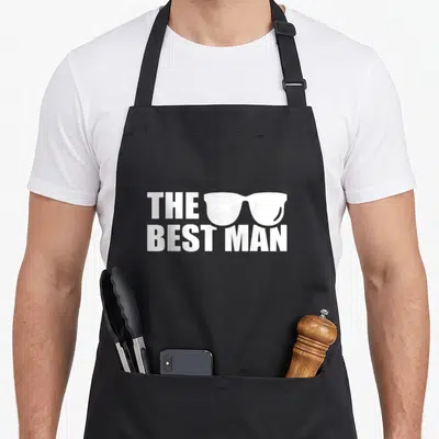 Stag bachelor party apron - White text THE BEST MAN with sunglasses graphic on grey background — shown on a black cotton canvas apron against a white background
