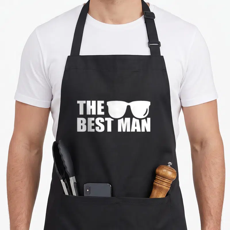 Stag bachelor party apron - White text THE BEST MAN with sunglasses graphic on grey background — shown on a black cotton canvas apron against a white background