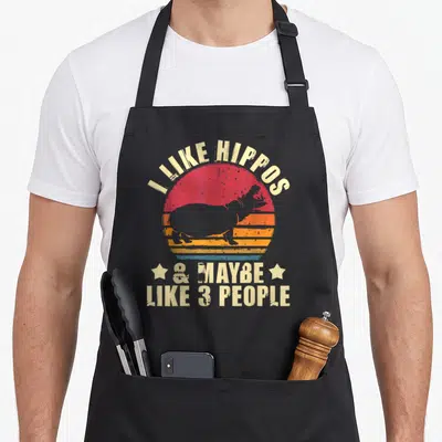Maybe hippo lover apron - I LIKE HIPPOS text over striped sun with hippo silhouette and two stars — shown on a black cotton canvas apron against a white background