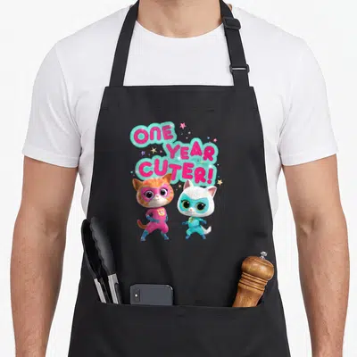 Cuter junior apron - Two superhero cats standing below stacked pink text reading ONE YEAR CUTER! with scattered stars — shown on a black cotton canvas apron against a white background