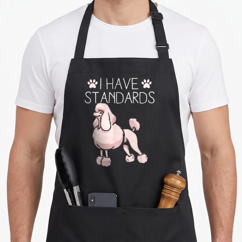 Retro dog standard size of apron - Pink cartoon poodle standing under white I Have Standards text flanked by two paw — shown on a black cotton canvas apron against a white background
