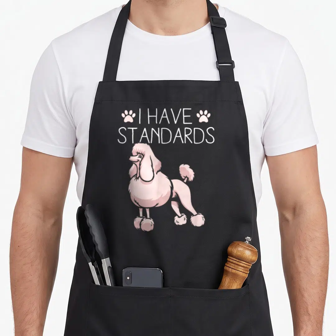 Retro dog standard size of apron - Pink cartoon poodle standing under white I Have Standards text flanked by two paw — shown on a black cotton canvas apron against a white background