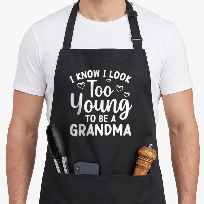 Young grandma apron - White stacked text I KNOW I LOOK Too Young TO BE A GRANDMA with four small hearts — shown on a black cotton canvas apron against a white background