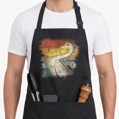 Zookeeper pelican bird apron - Retro pelican illustration with yellow beak over distressed red orange beige blue — shown on a black cotton canvas apron against a white background