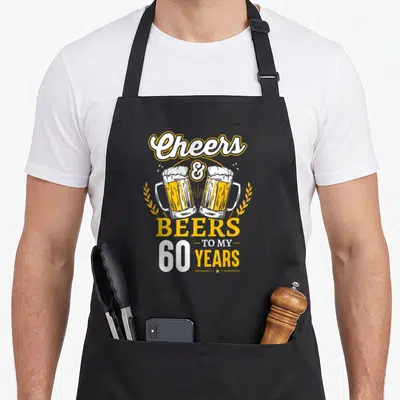 60th beer apron - Clinking beer mugs with wheat stalks and vertical text reading Cheers & Beers To My 60 Years — shown on a black cotton canvas apron against a white background