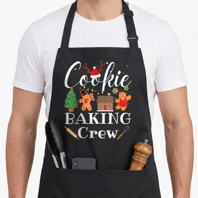 Crew christmas cookie baking team aprons - Gingerbread cookies, Santa hat, lights, rolling pin, whisk, and text — shown on a black cotton canvas apron against a white background