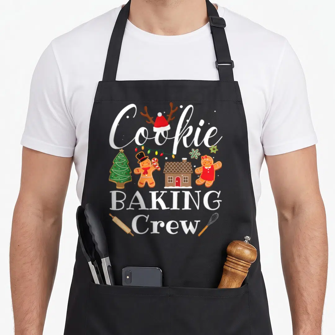 Crew christmas cookie baking team aprons - Gingerbread cookies, Santa hat, lights, rolling pin, whisk, and text — shown on a black cotton canvas apron against a white background