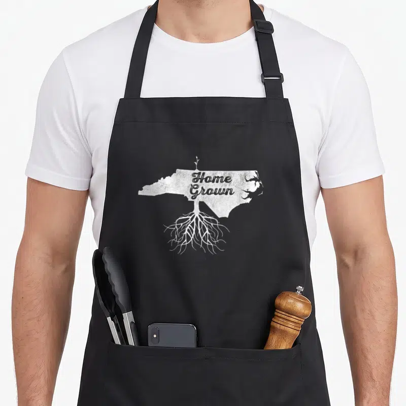 Carolina grown pride apron - White distressed NC map with roots, sprout, and centered Home Grown text — shown on a black cotton canvas apron against a white background
